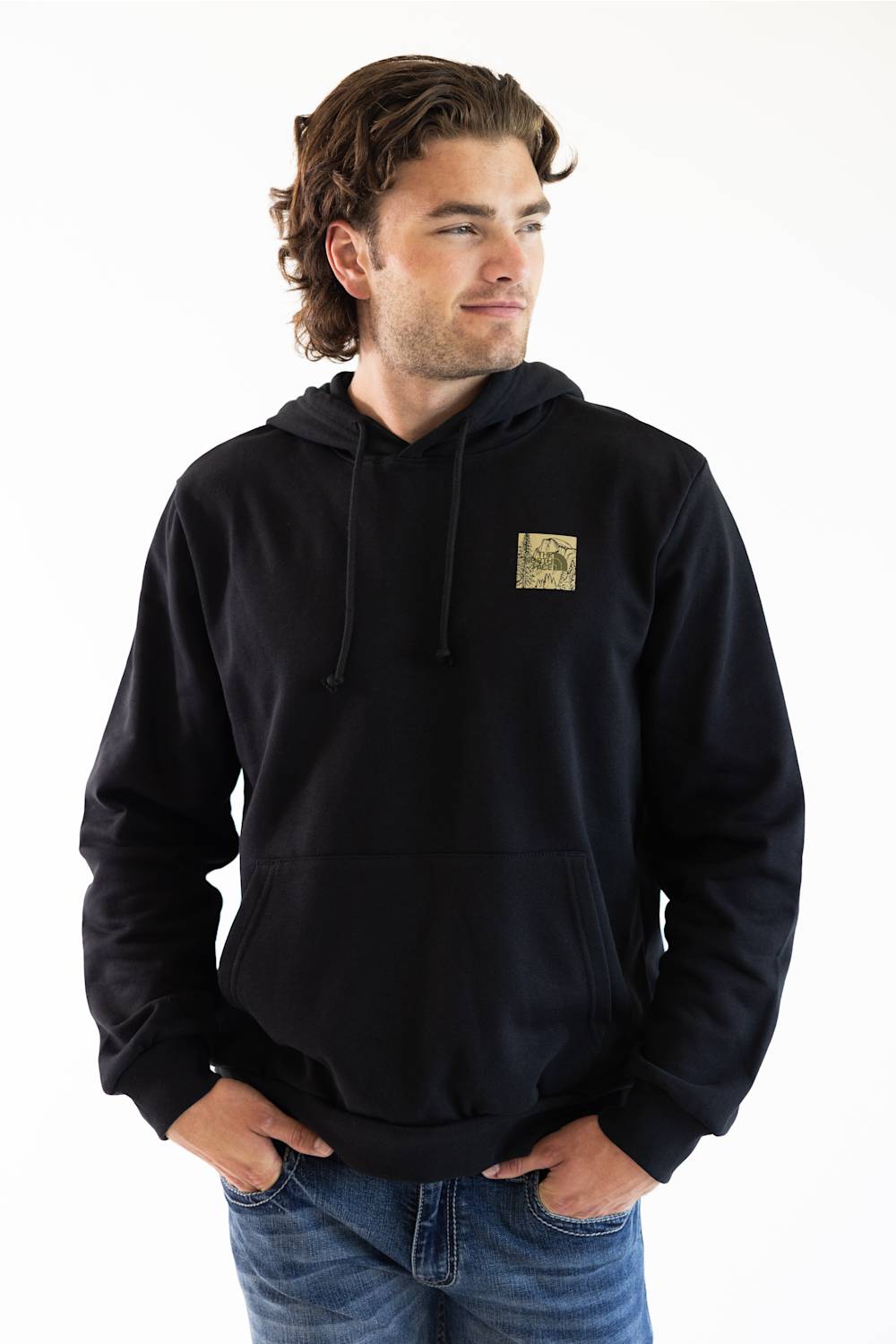 A man wearing a black pullover hoodie with a small square graphic patch on the left chest, paired with blue jeans, photographed against a plain white background.