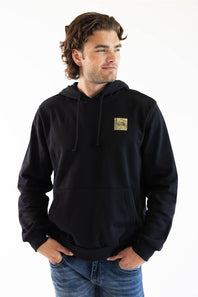 A man wearing a black pullover hoodie with a small square graphic patch on the left chest, paired with blue jeans, photographed against a plain white background.