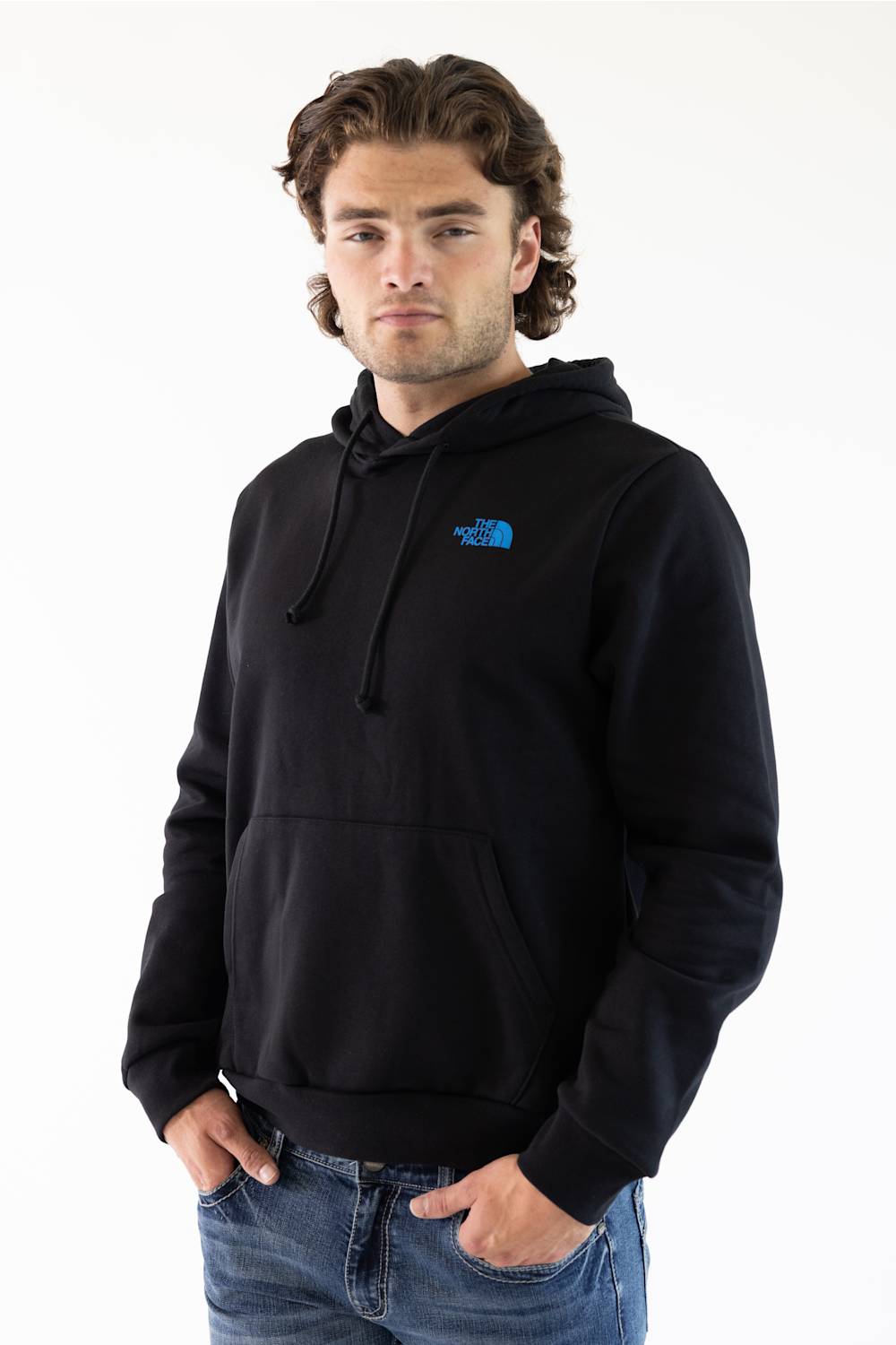A man wearing a black pullover hoodie with a small blue The North Face logo on the left chest, paired with blue jeans, against a plain white background.