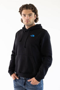 A man wearing a black pullover hoodie with a small blue The North Face logo on the left chest, paired with blue jeans, against a plain white background.