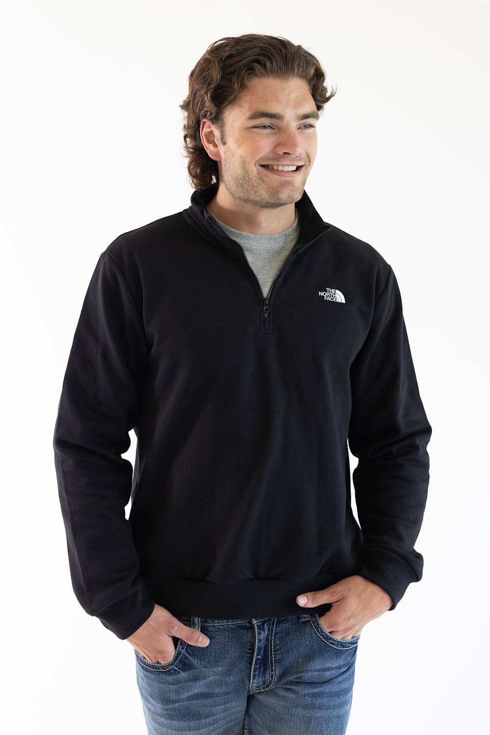 A man wearing a black long-sleeve quarter-zip pullover jacket and blue jeans, set against a plain white studio background.