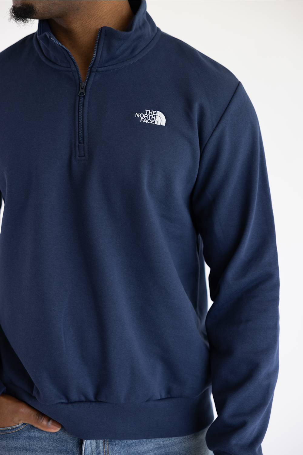 A man wearing a navy long-sleeve quarter-zip pullover jacket and blue jeans, set against a plain white studio background.