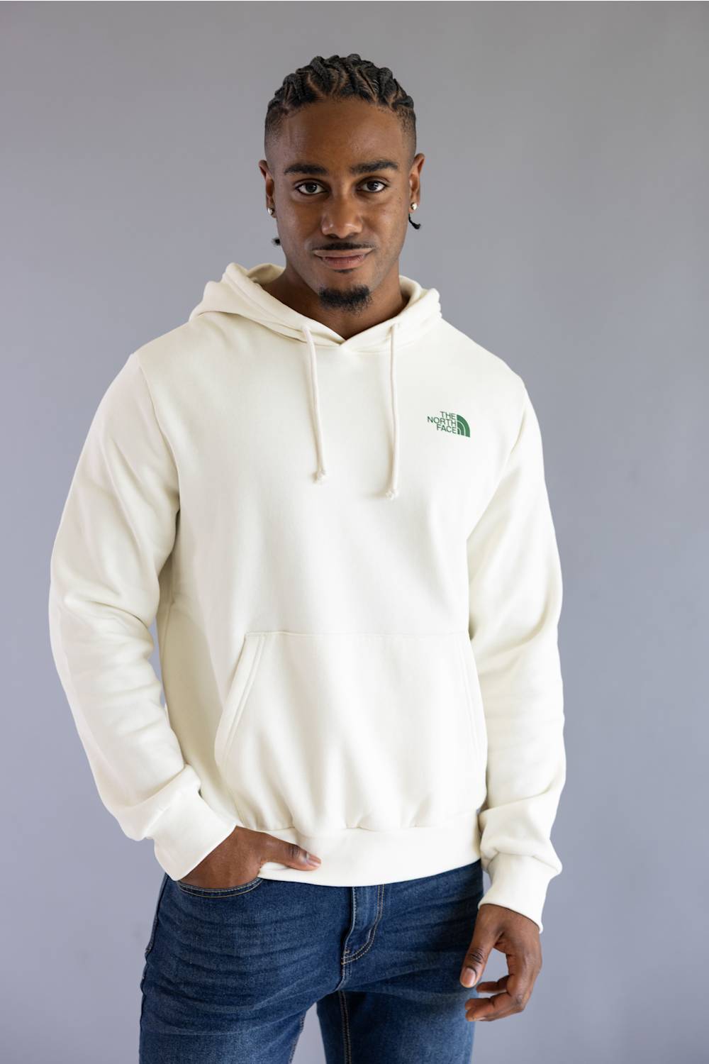 A man wearing an off-white hooded sweatshirt with a small green embroidered logo on the left chest, paired with blue jeans, standing against a solid gray studio background.