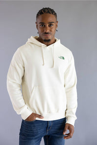 A man wearing an off-white hooded sweatshirt with a small green embroidered logo on the left chest, paired with blue jeans, standing against a solid gray studio background.