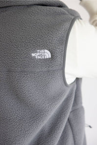 Close-up of a dark gray fleece vest with a white embroidered The North Face logo on the chest, shown against a white background.
