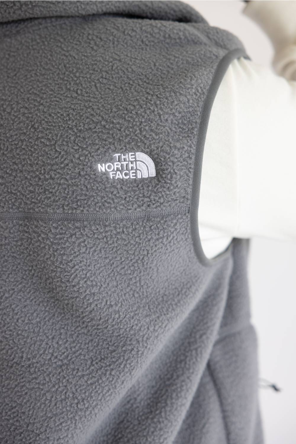 Close-up of a dark gray fleece vest with a white embroidered The North Face logo on the chest, shown against a white background.