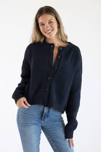 Front view of a female model wearing the Things Between Alyssa Cardigan in Navy, featuring a crew neckline, large front buttons, ribbed hem, and long sleeves paired with blue jeans.