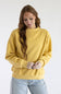 A woman wearing a mustard yellow crewneck sweatshirt and light blue jeans, standing against a plain white studio background.