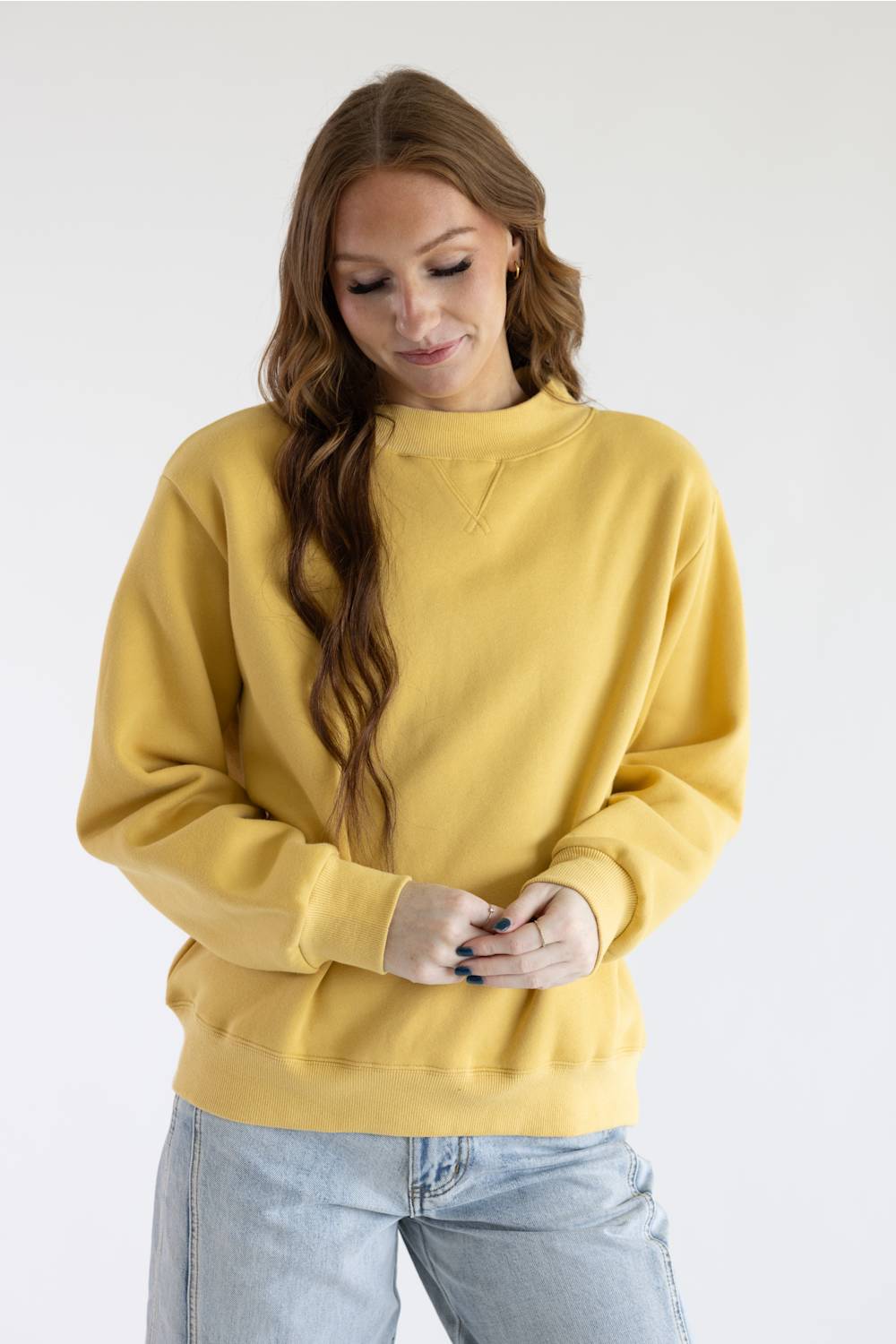 A woman wearing a mustard yellow crewneck sweatshirt and light blue jeans, standing against a plain white studio background.