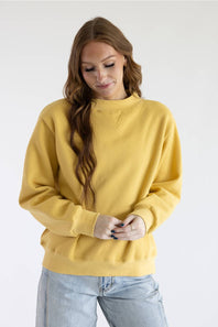 A woman wearing a mustard yellow crewneck sweatshirt and light blue jeans, standing against a plain white studio background.