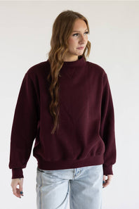 A woman with red hair wearing a loose-fitting, long-sleeve crewneck sweater in deep burgundy and blue jeans, standing against a plain white background.