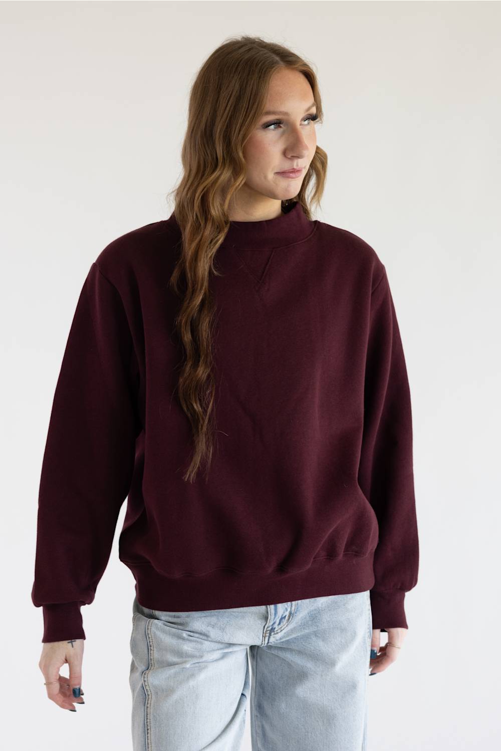 A woman with red hair wearing a loose-fitting, long-sleeve crewneck sweater in deep burgundy and blue jeans, standing against a plain white background.
