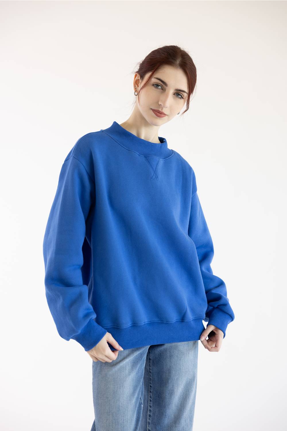A woman wearing a vibrant royal blue crewneck sweatshirt and light wash jeans, standing against a plain white studio background.