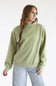 A woman wearing a sage green mock neck sweatshirt and light blue jeans, standing against a plain white studio background.