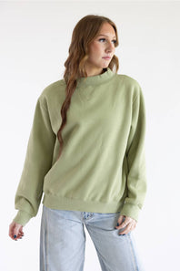 A woman wearing a sage green mock neck sweatshirt and light blue jeans, standing against a plain white studio background.