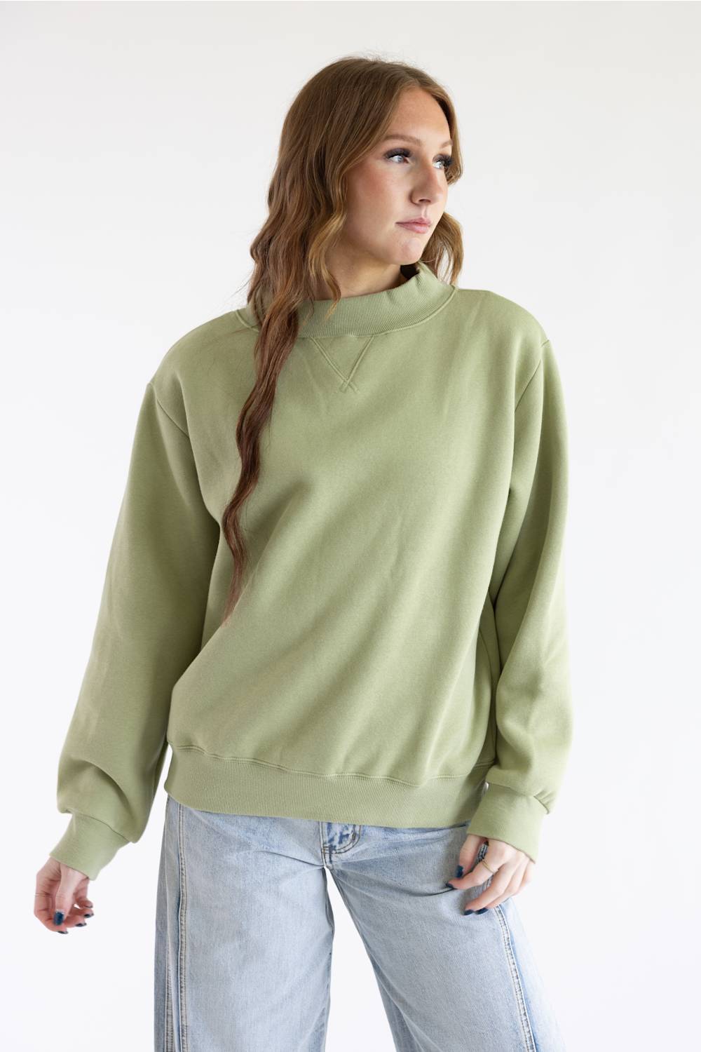 A woman wearing a sage green mock neck sweatshirt and light blue jeans, standing against a plain white studio background.