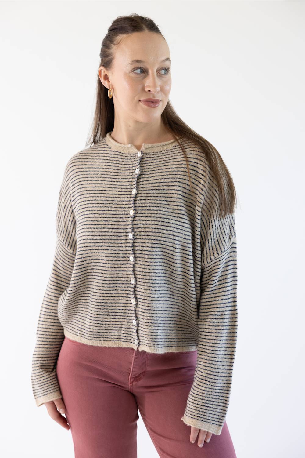 A woman modeling a cropped, long-sleeve knit cardigan with horizontal stripes in beige, featuring a button-down front. She is wearing dusty rose pants against a plain white studio background.