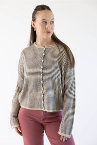 A woman modeling a cropped, long-sleeve knit cardigan with horizontal stripes in beige, featuring a button-down front. She is wearing dusty rose pants against a plain white studio background.