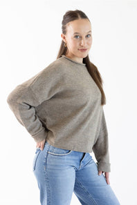 A woman modeling a cropped, long-sleeve knit sweater in a heathered taupe-brown color, paired with light-wash blue jeans, against a plain white studio background.
