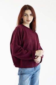 A woman with red hair wearing a loose-fitting, long-sleeve crewneck sweater in deep burgundy and blue jeans, standing against a plain white background.