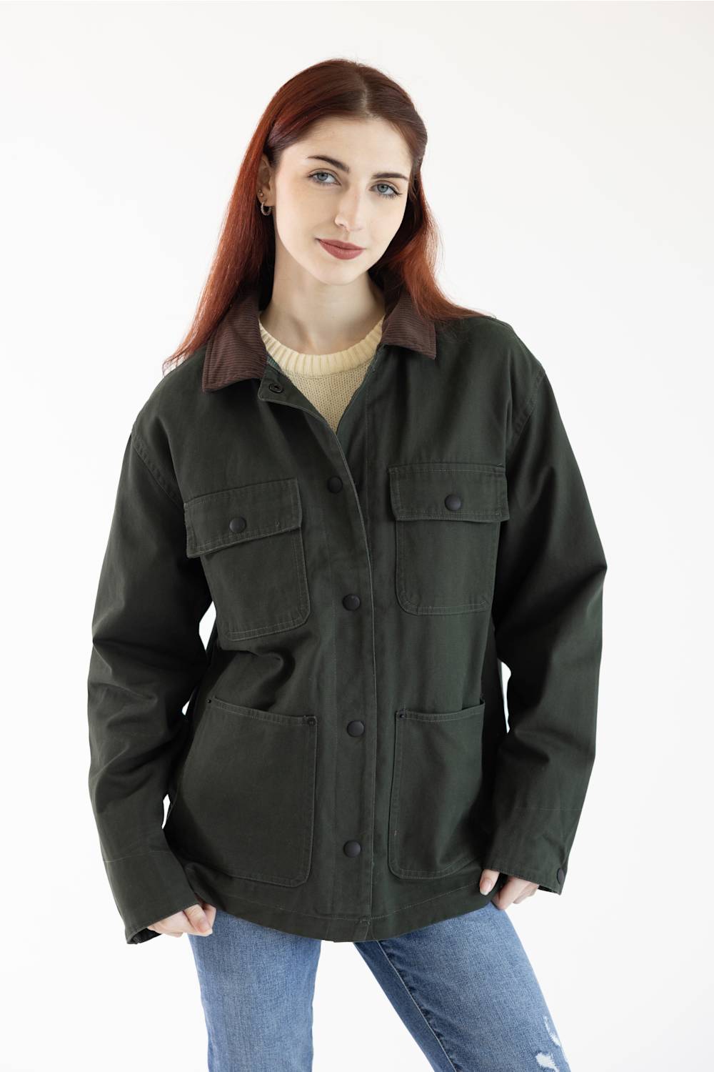 A woman with red hair wearing a dark olive green utility jacket with brown corduroy collar, a beige ribbed sweater, and blue jeans, photographed against a plain white studio background.