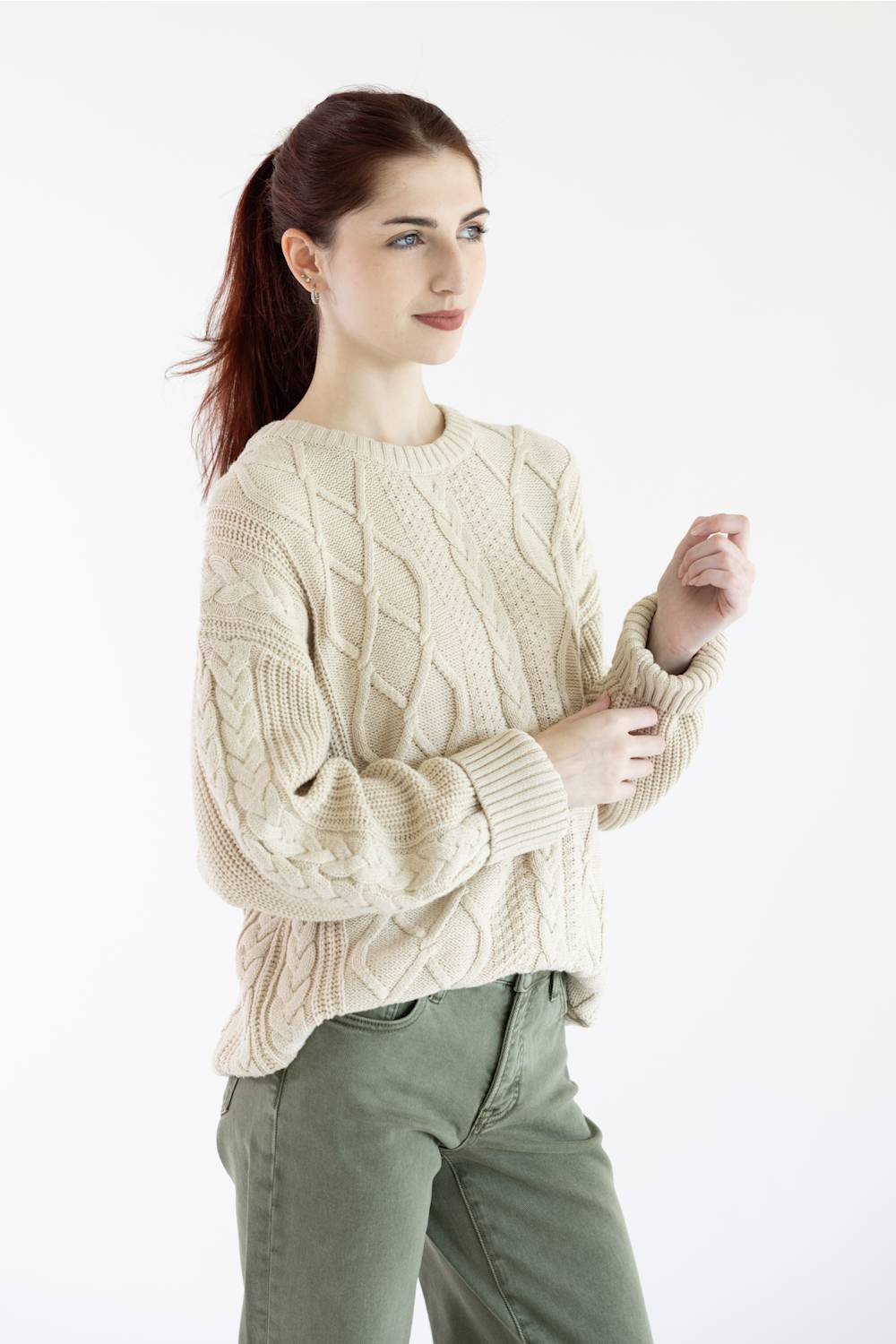 A woman wearing a cream cable-knit crewneck sweater and olive green pants, standing against a plain white studio background.