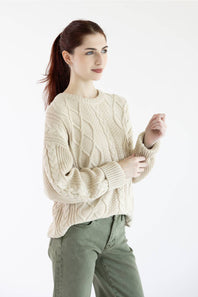 A woman wearing a cream cable-knit crewneck sweater and olive green pants, standing against a plain white studio background.