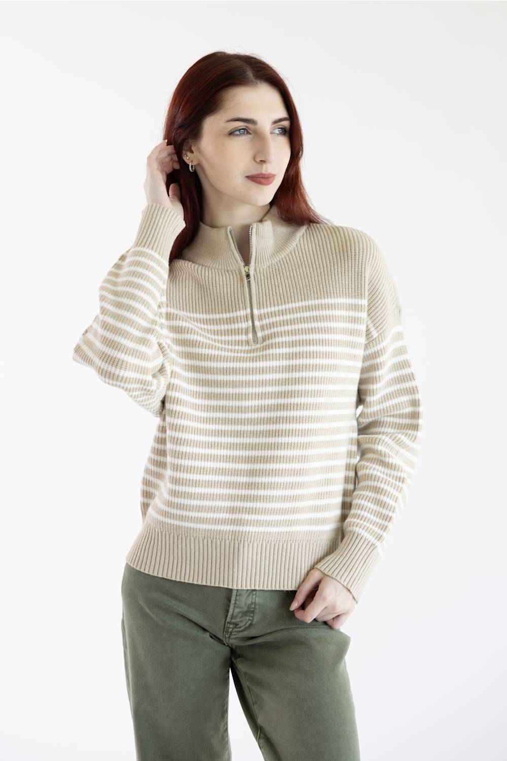 A woman with red hair wearing a beige and white horizontally striped quarter-zip sweater and olive green pants, standing against a plain white studio background.