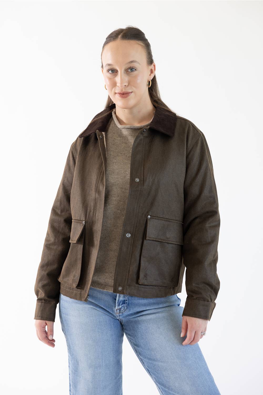 A woman modeling a dark brown suede jacket with a dark brown corduroy collar, worn over a brown knit sweater and blue jeans, against a plain white studio background.