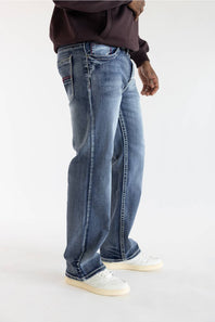 A person wearing blue denim jeans with a faded wash, styled with a dark brown hoodie and white sneakers, shown from the waist down against a plain white studio background.