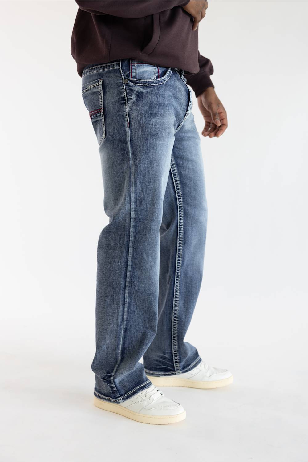 A person wearing blue denim jeans with a faded wash, styled with a dark brown hoodie and white sneakers, shown from the waist down against a plain white studio background.