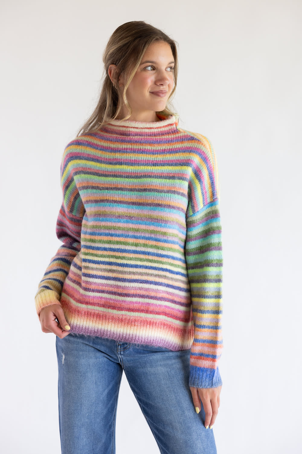 Woman wearing a striped multi rainbow colored mock neck sweater with blue denim jeans standing in front of a clean white background