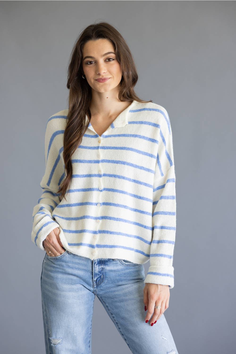 A woman modeling a cream-colored knit sweater with thin blue horizontal stripes, paired with light-wash blue jeans, against a solid gray studio background.