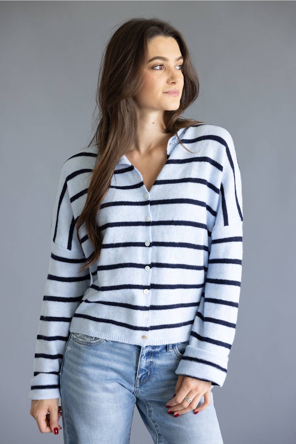 A woman wearing a white knit cardigan with navy blue horizontal stripes, paired with light-wash blue jeans, photographed against a solid gray studio background.