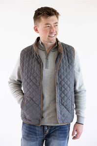 A male model wearing the FX Fusion Upland Quilted Field Vest in charcoal over a grey mock-neck quarter-zip sweater and blue jeans, turned slightly to the side with a smiling expression.
