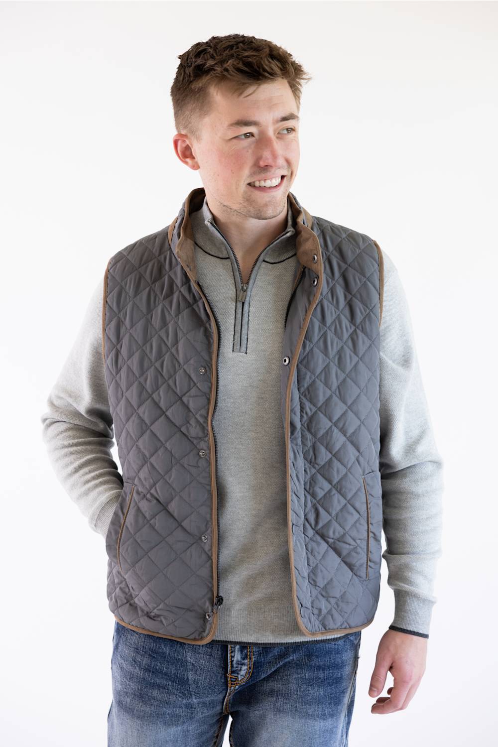 A male model wearing the FX Fusion Upland Quilted Field Vest in charcoal over a grey mock-neck quarter-zip sweater and blue jeans, turned slightly to the side with a smiling expression.