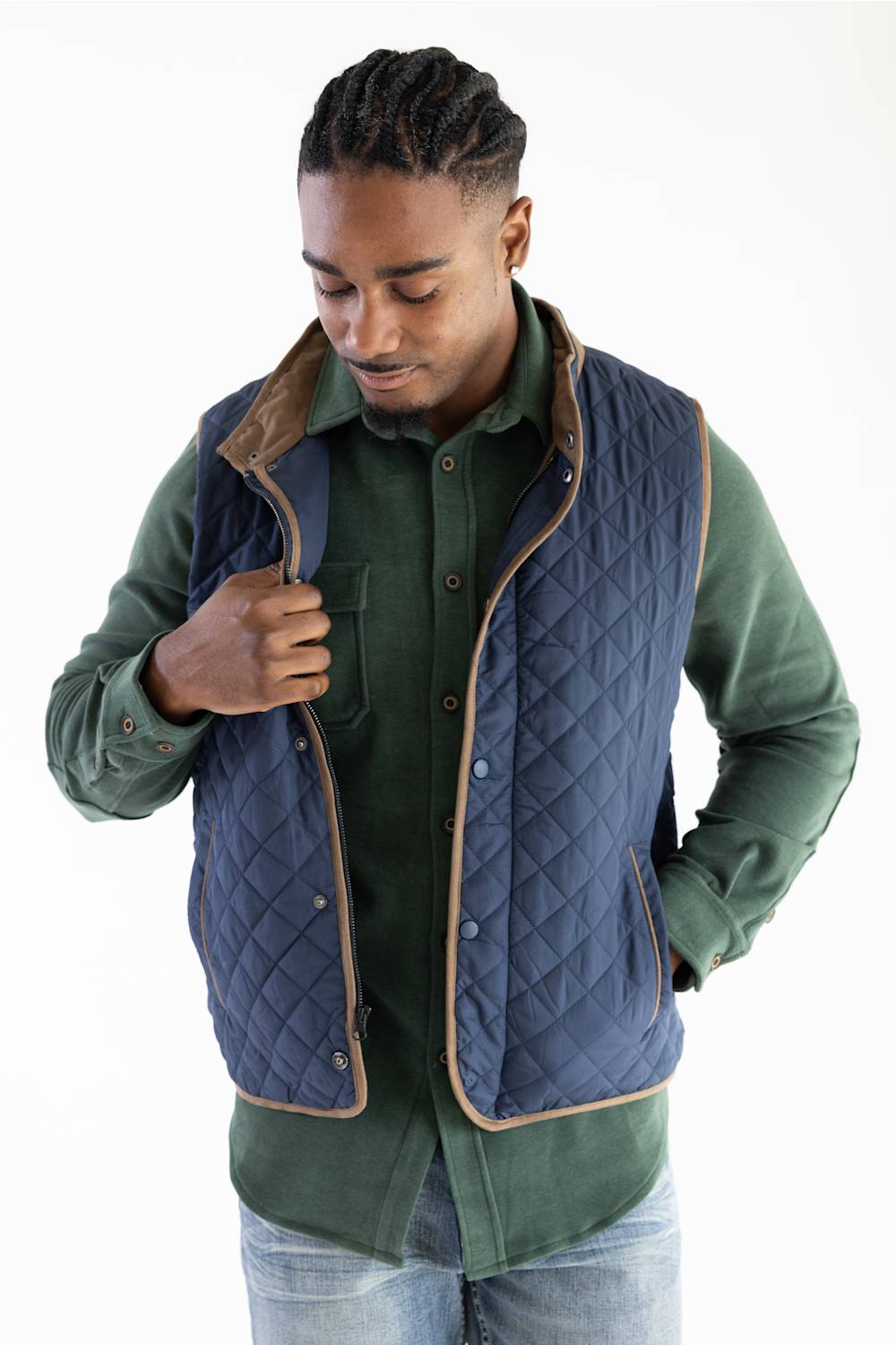 A man wearing a navy blue quilted vest with tan trim over a green button-up shirt and light blue jeans, photographed against a plain white studio background.