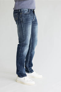 A person wearing blue denim jeans with a faded wash, paired with white sneakers and a light purple ribbed top, shown from the waist down against a plain light gray studio background.