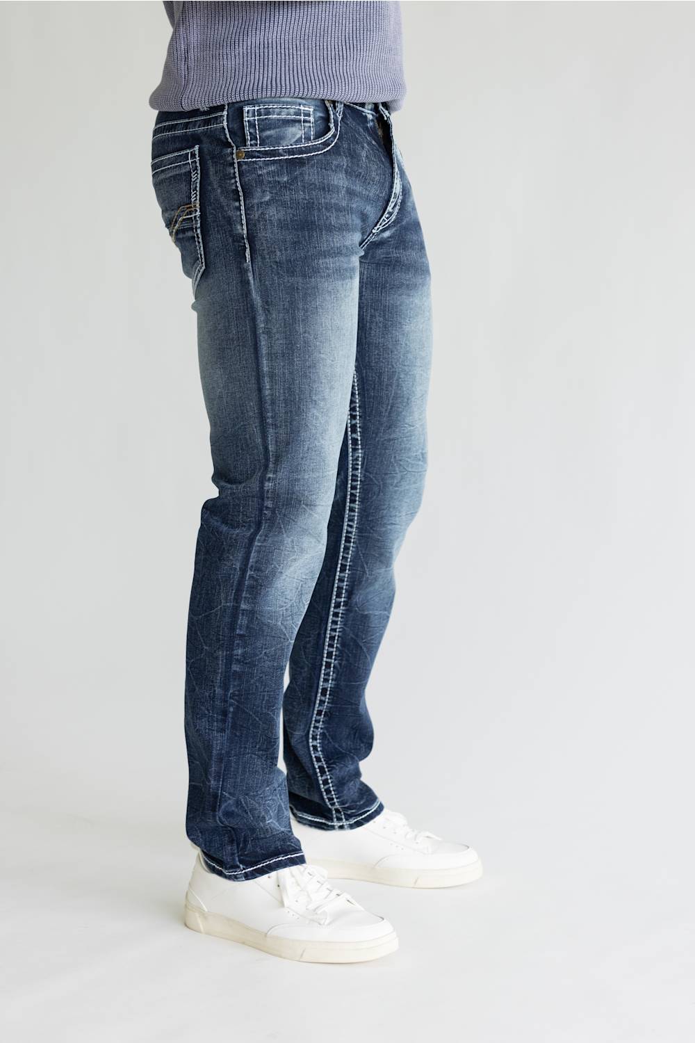 A person wearing blue denim jeans with a faded wash, paired with white sneakers and a light purple ribbed top, shown from the waist down against a plain light gray studio background.