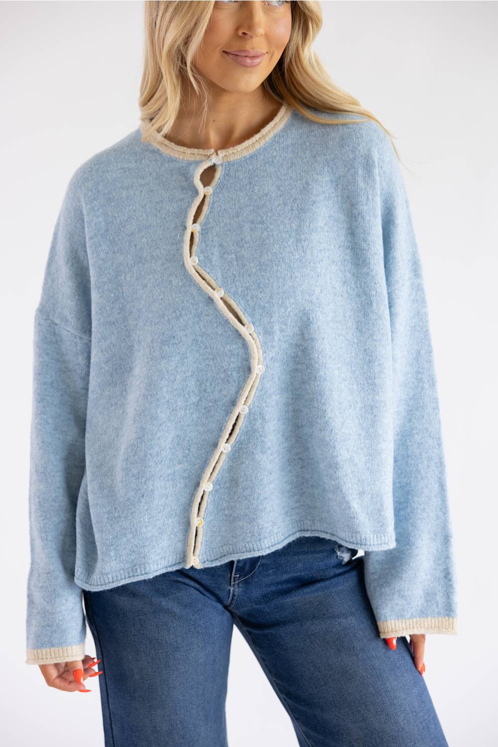 A woman wearing a light blue knit sweater with a zigzag beige trim detail down the front, paired with blue jeans, against a plain white background.