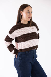 A woman wearing a brown and light pink striped knit sweater with dark blue jeans, photographed against a plain white studio background.