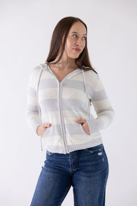 A woman wearing a white and light gray striped zip-up hoodie with drawstrings and front pockets, paired with blue jeans, photographed against a plain light gray studio background.