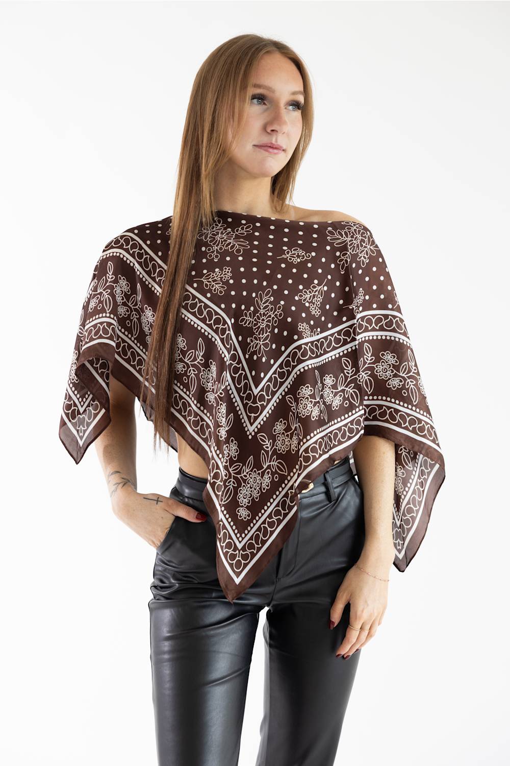 A woman modeling a brown and white patterned poncho top with black faux-leather pants, photographed against a plain white studio background.