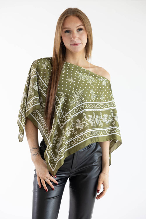 Front view of a model wearing an olive green asymmetric scarf poncho top with a white floral and polka dot print, styled off one shoulder with black leather pants against a plain white background.