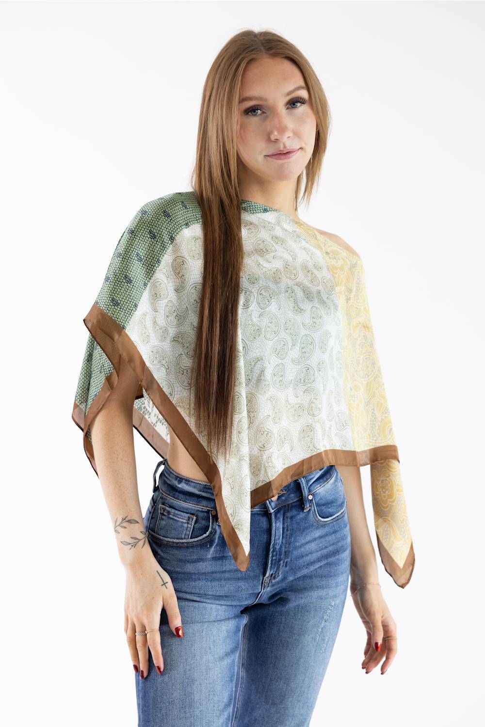 Medium close-up front view of a model with long straight brown hair wearing an asymmetric patchwork scarf poncho top featuring green, beige paisley, and yellow floral patterns with brown trim, styled off one shoulder with blue jeans against a plain white studio background.