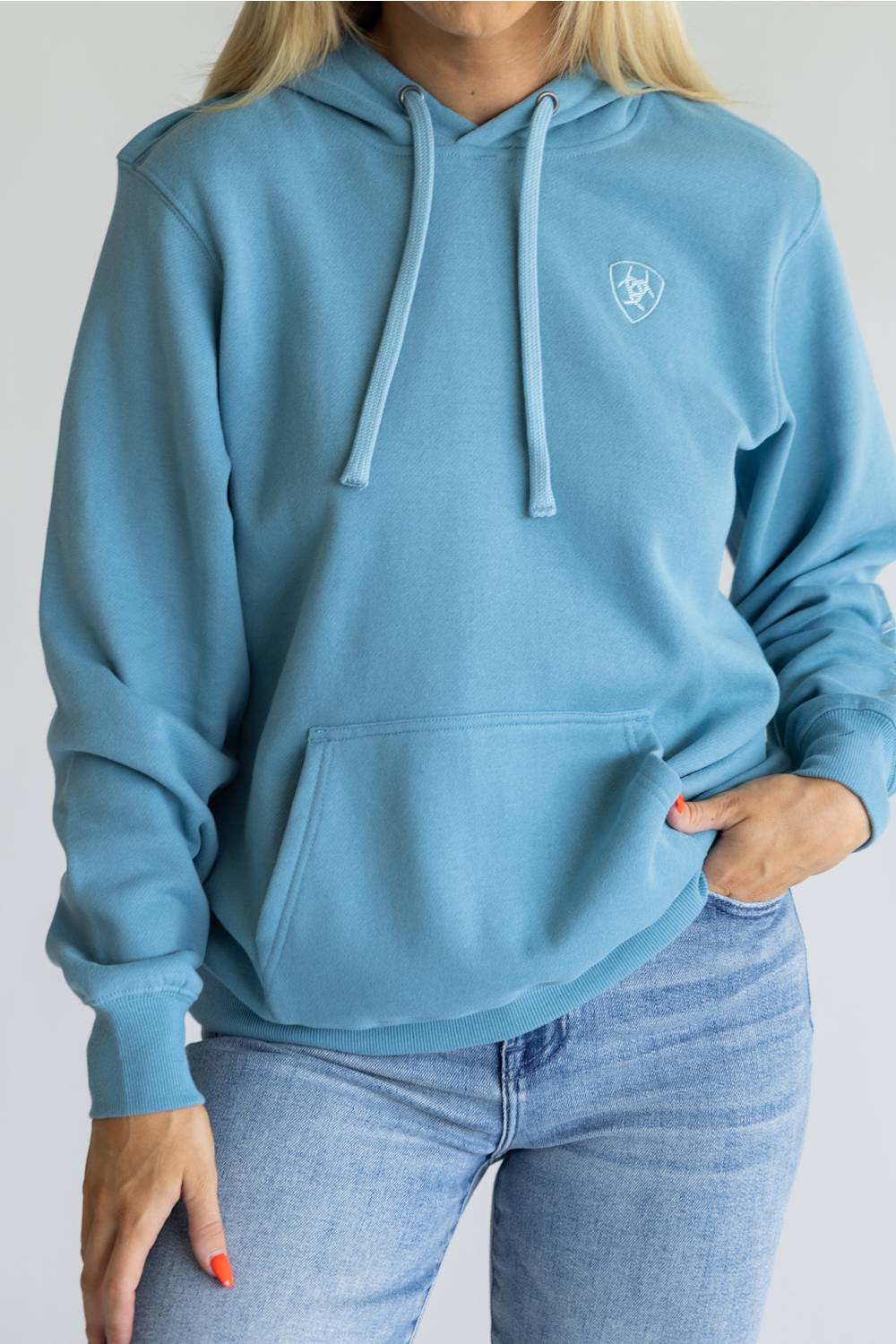A woman wearing a light blue pullover hoodie with a small white shield logo on the left chest, paired with light-wash blue jeans, shown against a plain light gray background.