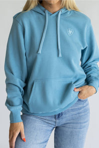 A woman wearing a light blue pullover hoodie with a small white shield logo on the left chest, paired with light-wash blue jeans, shown against a plain light gray background.