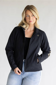 A blonde woman wearing a black zip-up jacket with a small shield logo on the left chest and gold stripes on the sleeve cuffs, paired with a black top and light blue jeans, against a light gray studio background.