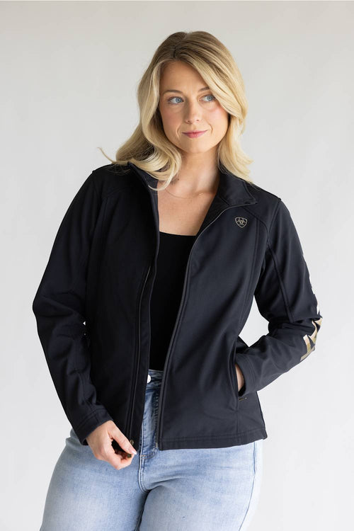 A blonde woman wearing a black zip-up jacket with a small shield logo on the left chest and gold stripes on the sleeve cuffs, paired with a black top and light blue jeans, against a light gray studio background.