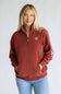 A woman wearing a rust-colored quarter-zip sweatshirt with a small shield logo on the left chest, paired with light blue jeans, standing against a light gray studio background.